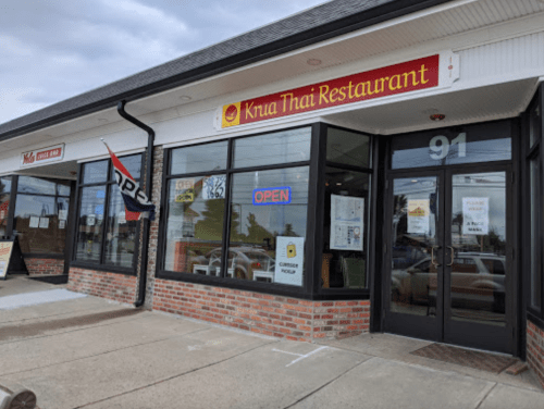 Exterior of Kua Thai Restaurant with a red and yellow sign, large windows, and an "OPEN" sign displayed.