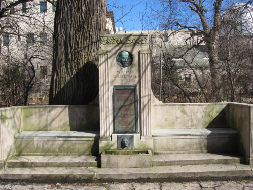 A stone monument dedicated to William Shakespeare, featuring a bust and an inscription, surrounded by trees.