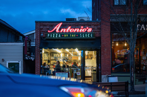 A cozy pizza shop, "Antonio's Pizza by the Slice," illuminated at dusk, with customers visible inside.