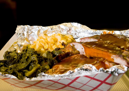 A foil-wrapped plate with barbecue meat, collard greens, and macaroni and cheese, served in a red-checkered container.