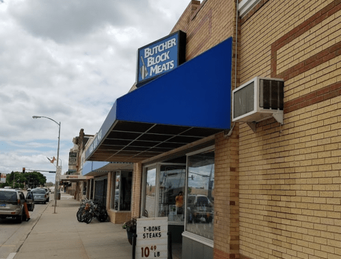 Exterior of Butcher Block Meats with a blue awning, showcasing a sign and a price for T-bone steaks.