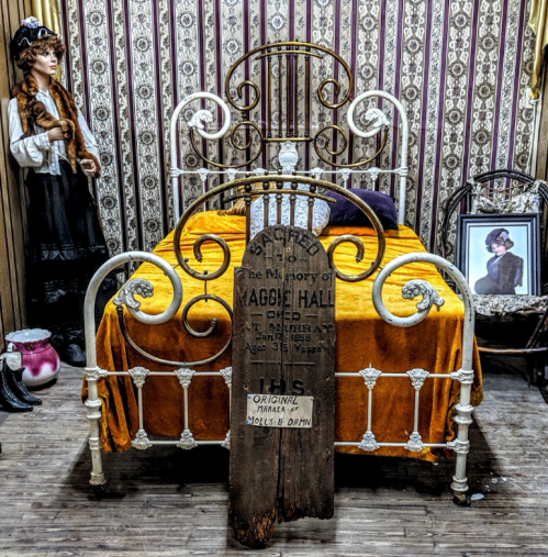 A vintage bedroom with an ornate bed, an orange coverlet, a historical plaque, and a woman in period clothing nearby.