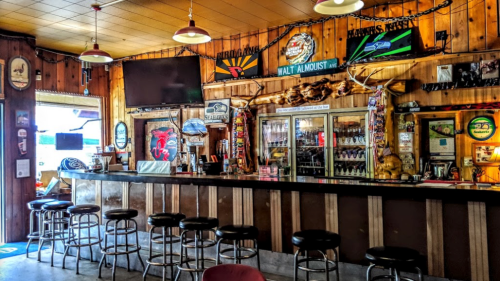 A rustic bar interior with wooden decor, stools, and various signs, featuring a large TV and a well-stocked drink display.