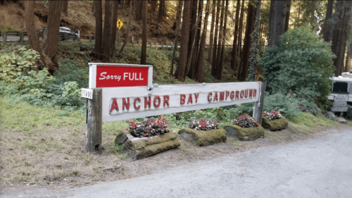 Sign for Anchor Bay Campground with "Sorry FULL" displayed, surrounded by trees and flowers along a gravel road.