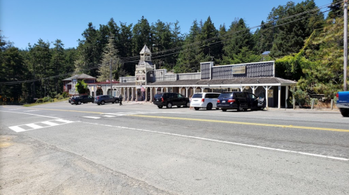 A roadside view of a building with a clock tower, surrounded by trees and parked cars on a sunny day.