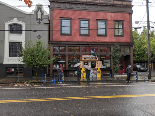 A red and gray building with people standing outside under umbrellas, and blue bike racks in front on a rainy day.