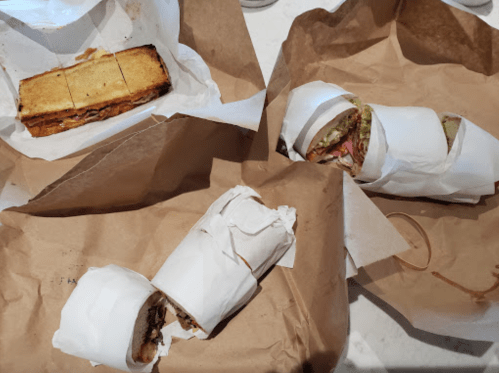Three wrapped sandwiches on crumpled brown paper, with one sandwich cut in half, showcasing various fillings.