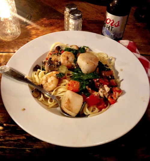 A plate of pasta with shrimp, scallops, vegetables, and a beer bottle in the background, set on a rustic table.