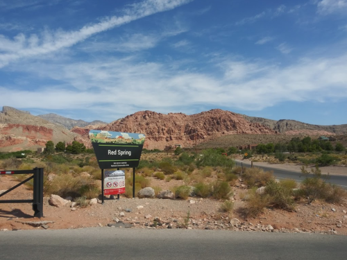 Sign for Red Spring with red rock formations and blue sky in the background, surrounded by desert vegetation.