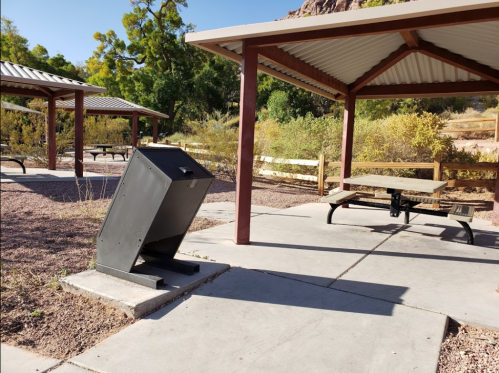 A metal trash bin near a picnic area with shaded tables and surrounding greenery.