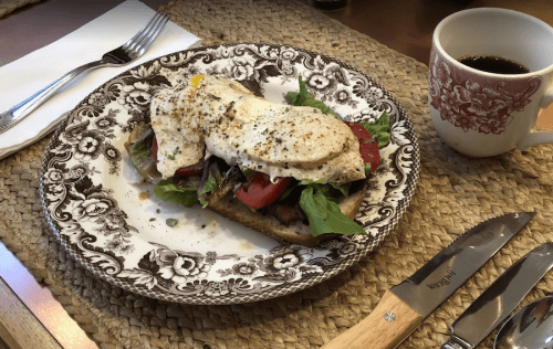 A plate with a toasted sandwich topped with greens, tomatoes, and a fried egg, alongside a cup of coffee.