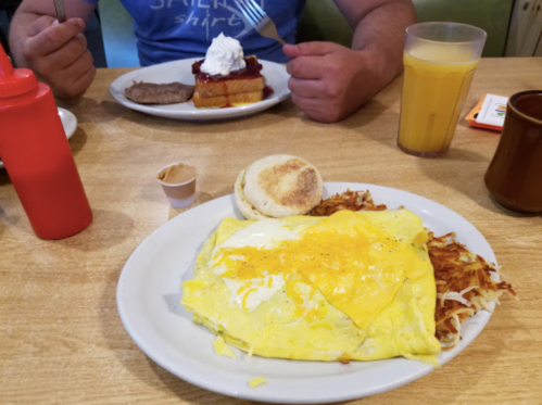 A plate of eggs, hash browns, and a muffin, with a person holding a fork in the background and a drink on the table.