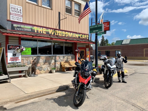 A motorcyclist stands next to two bikes in front of The Woodsman Cafe, featuring a rustic exterior and an American flag.