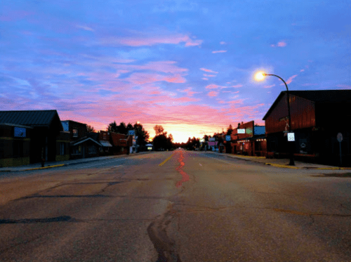 A quiet street at sunset, with colorful clouds and buildings lining the road, illuminated by a streetlamp.