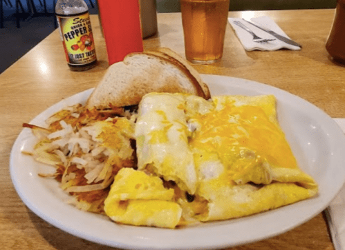 A plate of cheesy omelets with hash browns and toast, accompanied by a drink in a diner setting.