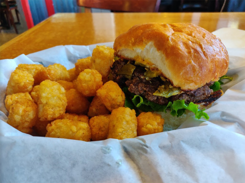 A juicy burger with lettuce and pickles, served with a side of crispy tater tots on a paper-lined plate.