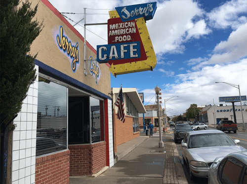 A colorful café sign for "Jerrys" hangs above a street lined with cars and shops under a partly cloudy sky.