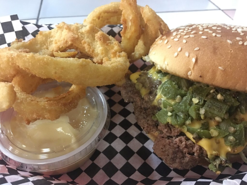 A sesame seed burger topped with cheese and jalapeños, served with onion rings and a side of creamy dip.