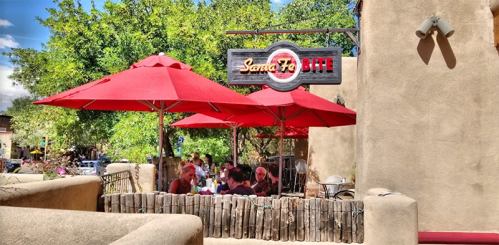 Outdoor dining area with red umbrellas at Santa Fe Bite, surrounded by greenery and a rustic wall.