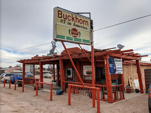 Exterior of Buckhorn restaurant with a sign promoting green chile cheese burgers, surrounded by cars and a cloudy sky.