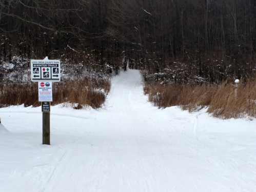 Snow-covered trail leading into a wooded area, with signs for winter trails on the left.