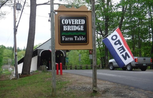 Sign for "Covered Bridge Farm Table" with a flag nearby, set in a wooded area with a covered bridge in the background.