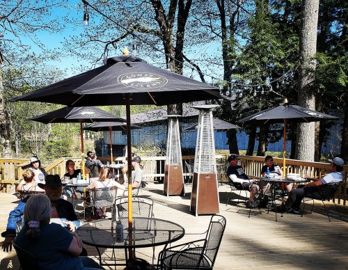 A lively outdoor deck with people sitting under umbrellas, enjoying drinks by a lake surrounded by trees.