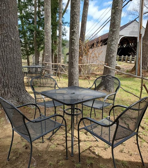 A black metal table surrounded by four chairs, set in a grassy area among trees with a rustic building in the background.