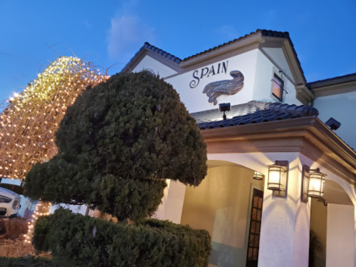 A building with "Spain" sign, featuring decorative lights and a neatly trimmed bush in the foreground at dusk.