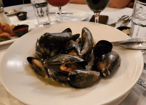 A bowl of mussels in a savory broth, served on a table with drinks and condiments in the background.