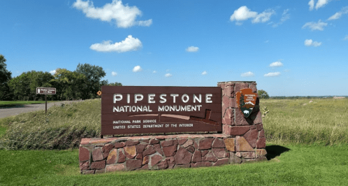 Sign for Pipestone National Monument, featuring stone structure and blue sky with clouds in the background.