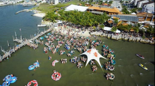 Aerial view of a crowded beach area with people on paddleboards and a large tent by the water, surrounded by palm trees.