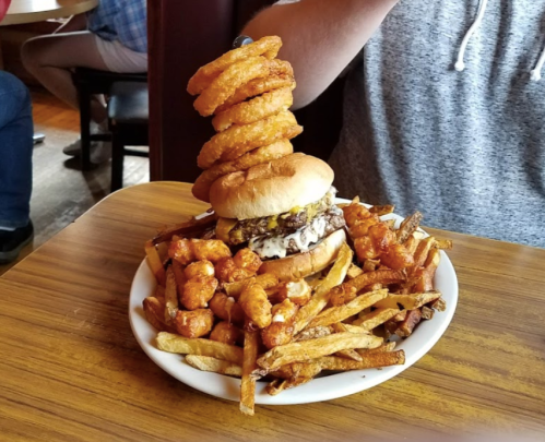 A towering burger topped with onion rings, surrounded by a mound of fries and crispy tater tots on a plate.