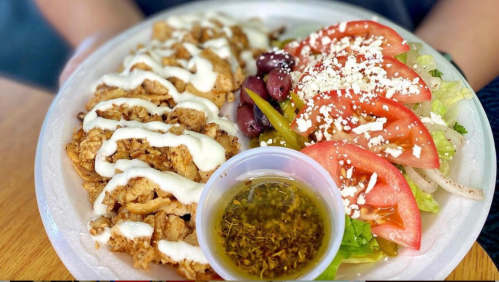 A plate with grilled chicken drizzled with sauce, a fresh salad with tomatoes, beans, and a small cup of dressing.
