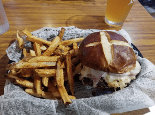 A pretzel bun burger with melted cheese and toppings, served with a side of crispy fries on a newspaper-lined plate.