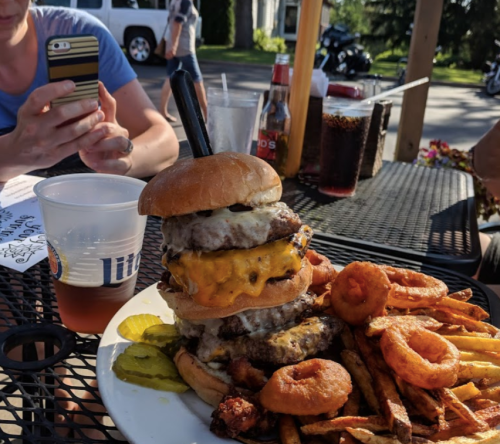 A towering burger with multiple patties and cheese, served with fries, onion rings, and pickles on a table.