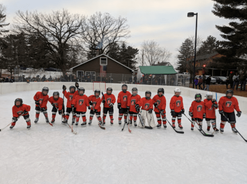 A youth hockey team in red jerseys poses on an outdoor rink, with trees and a building in the background.