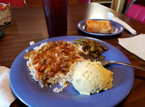 A plate of rice topped with a savory dish, green beans, mashed potatoes, a slice of toast, and a drink in the background.