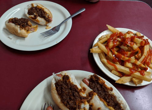 Two chili hot dogs on plates with a side of fries drizzled with ketchup on a red table.