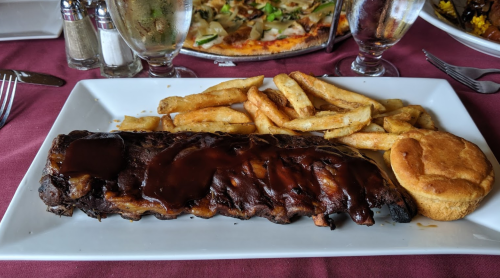 A plate with BBQ ribs, French fries, and a cornbread muffin on a maroon tablecloth.