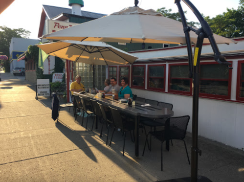 A group of three people sits at a long outdoor table under an umbrella, enjoying a meal at a restaurant.