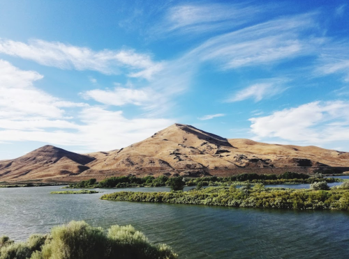 A serene landscape featuring a river, lush greenery, and a dry, rolling hill under a blue sky with scattered clouds.