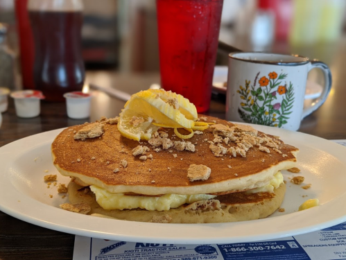 A large pancake topped with lemon slices and crushed cereal, served with a cup of coffee and a red drink.