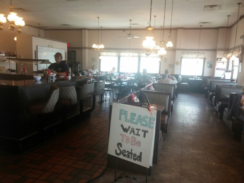 A spacious diner interior with booths, tables, and a "Please wait to be seated" sign at the entrance.