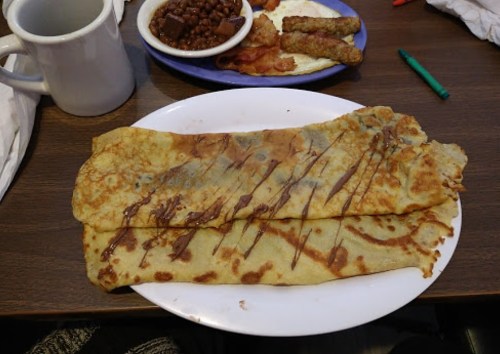 Two large crepes drizzled with chocolate sauce on a white plate, with a side of beans and breakfast items in the background.
