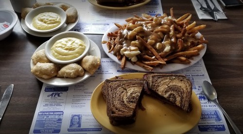 A table spread with poutine, fries, pierogies, and two slices of marbled rye bread on a menu-covered surface.