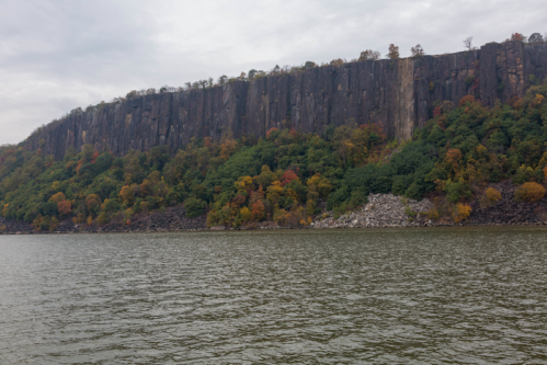 A rocky cliff covered in autumn foliage rises above a calm river under a cloudy sky.