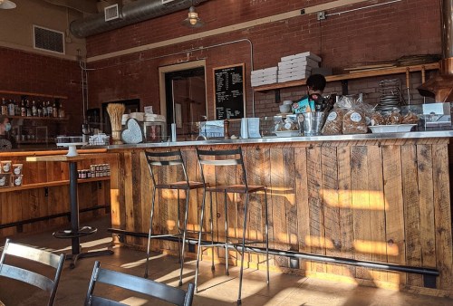 A cozy café interior featuring a wooden counter, bar stools, and a menu board, with a barista preparing drinks.