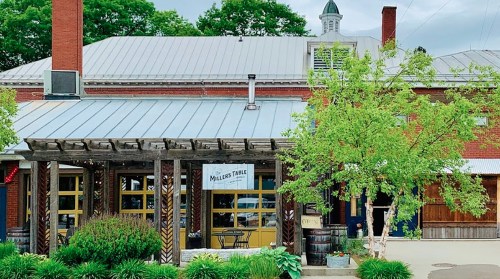 A rustic restaurant with a metal roof, wooden porch, and greenery, featuring a sign that reads "Millie's Table."