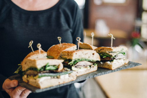 A person holds a slate platter with four stacked sandwiches, each topped with toothpicks and filled with greens and meats.